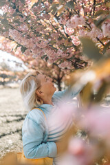 Beautiful happy blonde senior 60s woman enjoying sunny day in park during cherry blossom season on a nice spring day. 