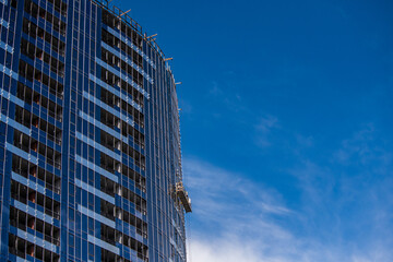 Building under construction against the blue sky on a sunny day, high-altitude work, a cradle on the wall of the house with workers installing glass