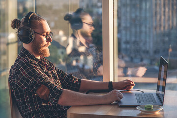 A man with a beard works at a computer, sitting in the office by the window.