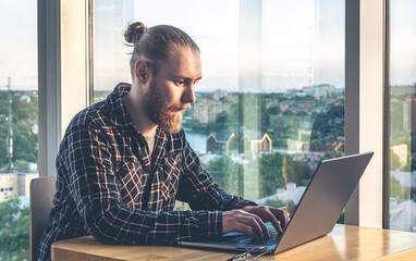 A man with a beard works at a computer, sitting in the office by the window.
