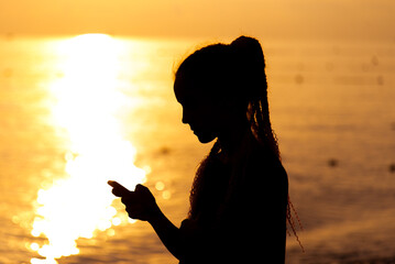 A teenage girl with a phone in the background of the sunset sea. Rest. Communication online. Summer.