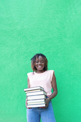 A smiling African-American woman holds a stack of books on a green background. Back to school. Copy space