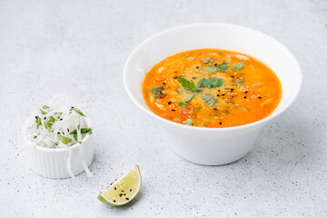 Orange color tom yum soup next to a cup with white noodles and lime slice