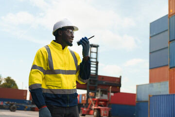 Foreman worker in uniform commanding workforce via walkie talkie at container yard outdoor. Black...