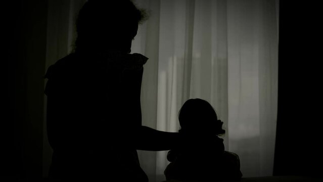 Bored child in home quarantine. Silhouette a little girl with a doll in protective medical masks sits on the bed and looks out the window. Allergy, covid-19 coronavirus pandemic, epidemic prevention.