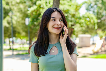 Happy woman wearing tee standing on city park, outdoors talking on mobile phone with friends or boyfriend with smiles.