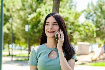 Cheerful brunette woman wearing tee standing on city park, outdoors talking on mobile phone with friends or boyfriend with smiles.