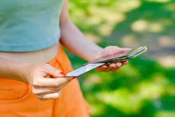 Young caucasian woman wearing tee standing on city park, outdoors holding mobile phone credit bank card do online shopping provide internet payment. Safe mobile banking.