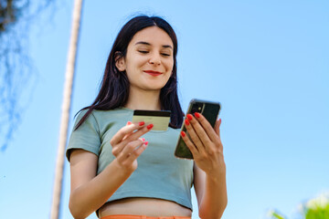 Young brunette girl smiling happy wearing turquoise t-shirt standing on city park, outdoors showing plastic credit or debit card while holding mobile phone. Online shopping, banking and transaction.