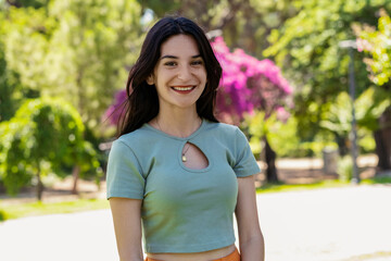 Young brunette girl smiling happy wearing turquoise t-shirt standing on city park, outdoors looking at camera while posing. Natural beauty concept. Beautiful girl. Pretty young woman.