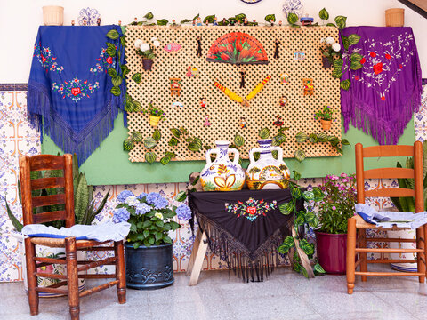 t&iacute;pico patio andaluz con sillas de Gea ,mesa con mantos de Manila, abanicos y botijo pata el agua y macetas colgadas en las pared. fiesta t&iacute;pica en los Corralones de M&aacute;laga