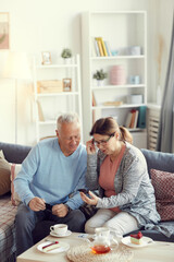 Happy mature woman in homewear sitting on sofa and watching old photographs while remembering past with senior husband