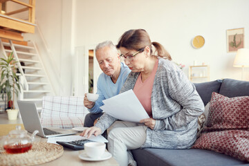 Serious senior woman in glasses sitting on sofa and looking at papers while analyzing home finances with husband