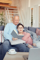Fototapeta na wymiar Content relaxed senior couple in casual clothing resting on sofa and reading book together