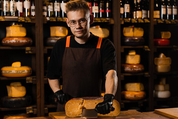 Handsome man worker of cheese shop cut limited gouda with knife. Cheese sommelier working in cheese...