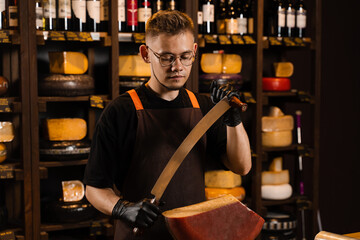 Handsome cheese sommelier with big knife for cut cheese. Bearded hipster in fashion glasses working in cheese shop. Creative idea for advertising.