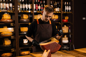 Handsome cheese sommelier with big knife for cut cheese. Bearded hipster in fashion glasses working in cheese shop. Creative idea for advertising.