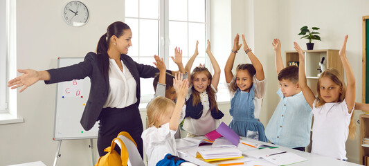 Cheerful teacher and students having fun in class. Happy young woman and little children standing around table with books in modern classroom, raising their hands up and smiling. Back to school banner