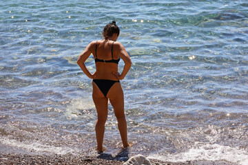 Woman in swimsuit going to swim in sea and tries the water temperature with her feet. Vacation on a pebble beach of summer resort with rocky bottom
