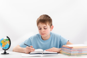 Boy of 8-9 years old does homework. Schoolboy sits at table with book, notebooks and globe. Isolated on white background.