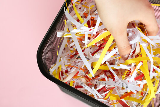 Woman putting shredded colorful paper strips into trash bin on pink background, closeup