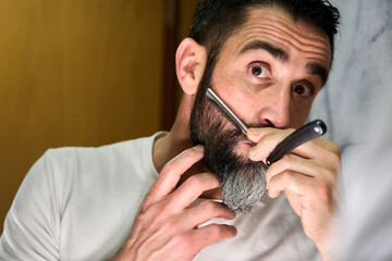 Young man with a beard shaving with a traditional knife in front of the mirror
