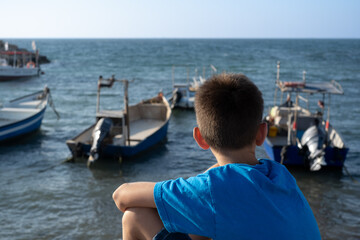 Fototapeta na wymiar The israeli teen boy sits and looks at the boats in the mediterranean sea. Dreams concept