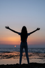 Fototapeta premium Teen woman looking to sunset, rejoicing by raising her hands up against the sea in israel. Triumph silhouette