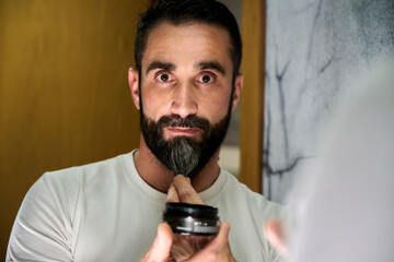 Young man applying wax and oil to his beard in front of mirror