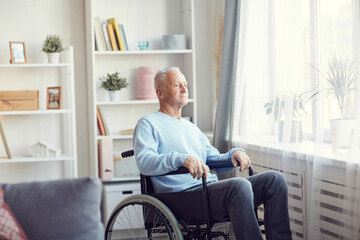 Content pensive senior man in casual sweater and jeans sitting in wheelchair and looking out window...