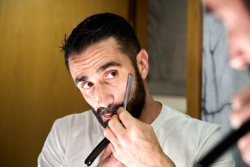 Young man with a beard shaving with a traditional knife in front of the mirror