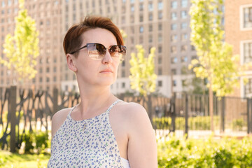 Portrait of a red-haired woman with a short haircut woman with sunglasses. A woman in off-the-shoulder clothes looks away against the backdrop of a multi-storey residential area