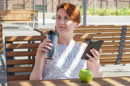 Woman Holds An Ebook Device And A Reusable Coffee Cup In Her Hands. A Woman Reads A Book Near The Office During A Work Break. Lunch Break On A Summer Day.