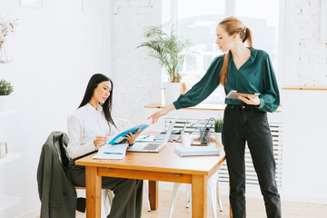 two young business women professionals in formal wear clothes work in modern office using laptop, tablet, brainstorm and search for solutions together, confident independent Asian girl solves problems