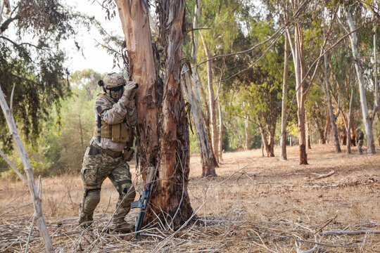 A full-length military man hid behind a tree and makes reconnaissance. A man in camouflage scouts the situation. Airsoft game
