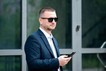 A business man in dark sunglasses with a phone in his hands stands outside the business building talks on the phone and looks at the watch. He is dressed in a light shirt and casual style jacket.