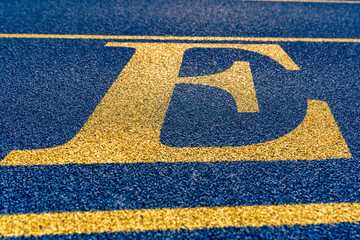 Close up the letter, initial E on a new blue running track with yellow lane lines.