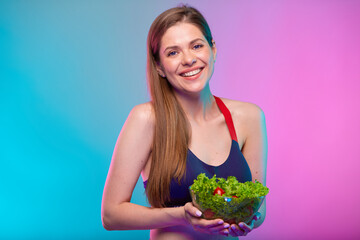 Smiling sporty woman in sportswear holding green salad in big glass bowl. Female fitness portrait isolated on neon color background.