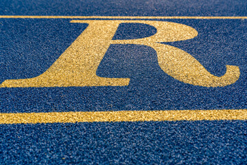 Close up the letter, initial R on a new blue running track with yellow lane lines.