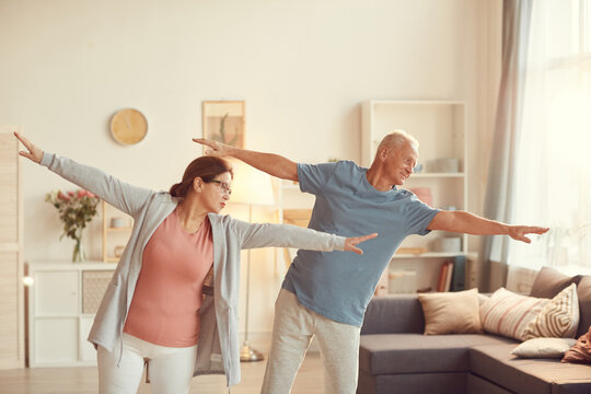 Content senior couple in activewear standing in living room and outstretching arms while training together at home
