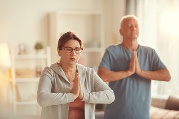 Fototapeta premium Serene aged couple concentrated on meditation standing in living room and making Namaste gesture