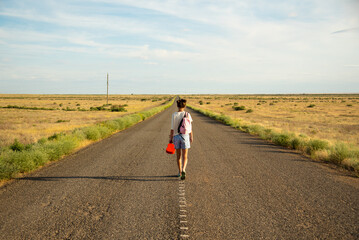 Obraz premium A girl walks along an empty road in the steppe. She has a ukulele in her hands and a backpack on her shoulders. It's summer and warm outside. Open road.