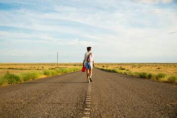 A girl walks along an empty road in the steppe. She has a ukulele in her hands and a backpack on...