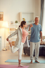 Fototapeta premium Careful handsome senior husband in tshirt holding hand of wife while assisting her to perform balance pose at home