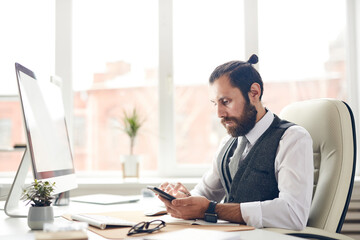 Serious young bearded businessman with ponytail sitting at desk with computer and viewing notes of tasks on smartphone