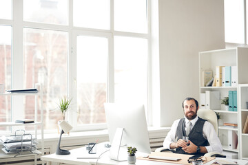 Portrait of cheerful successful young bearded businessman with beard sitting at table in spacious...