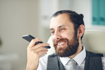 Positive handsome bearded businessman sitting on office chair and using smartphone while recording funny message for colleague
