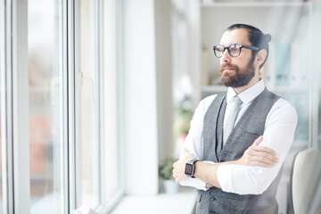 Fototapeta premium Serious purposeful young bearded businessman in gray waistcoat standing in modern office and looking out window