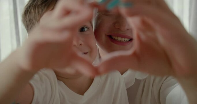 Look at camera laughing showing family love concept, close-up portrait. Affectionate happy young woman mom and cute adorable little child son make a heart shaped hand gesture.