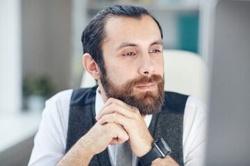 Close-up of concentrated young bearded man with wristwatch narrowing eyes while reading from computer screen